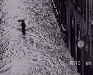 Una persona con l'ombrello percorre una strada invasa dalle acque. Immagine RAI
