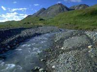 Il letto di un fiume in primo piano, con prati, montagne e cielo azzurro sullo sfondo